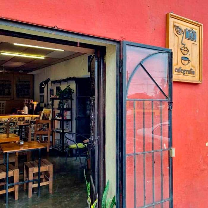 Exterior view of Cafegrafía in Antigua Guatemala, showing its red facade, open metal door, and wooden sign with a stylized face, coffee cup, and camera logo. The cozy interior features wooden tables, art displays, and a warm coffee shop ambiance.