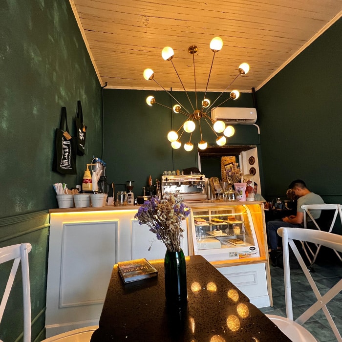 nterior of a small, modern coffee shop with minimalist decor, soft warm lighting, and a cozy seating area. A single table is set beside a neutral-toned wall with a large, artistic plant arrangement, capturing a calm and intimate atmosphere.