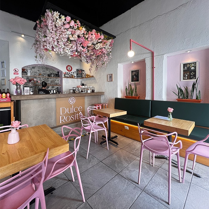 Cozy café interior at Dulce Rosita in Antigua Guatemala, featuring pink floral décor hanging from the ceiling, wooden tables with pink chairs, and a modern counter with desserts and drinks.
